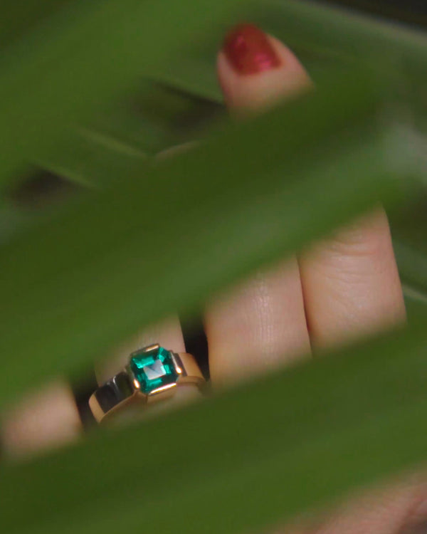 Hand behind palm leaf fronds wearing an emerald ring form the Fold Collection