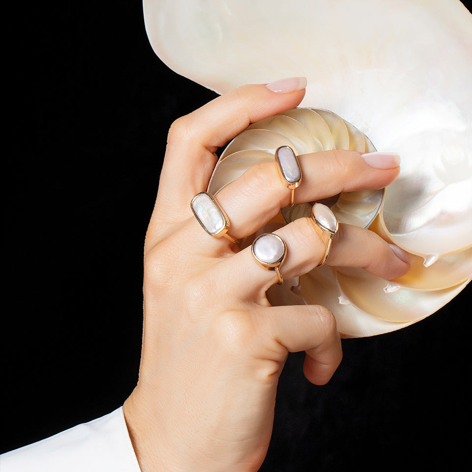 Gifts for Yourself Collection full bezel rings shown on a model's hand that is holding a large white seashell, on a neutral-light background.