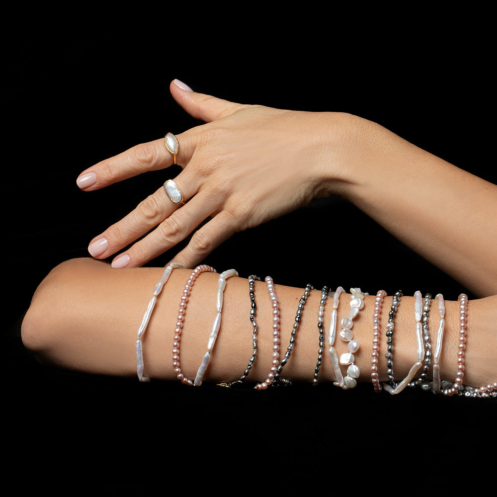 Photo of arm and hand wearing pearl bracelets and rings on a black background. 