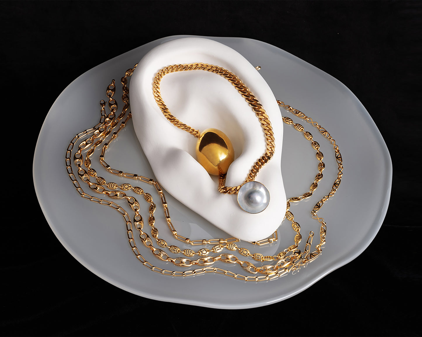 Styled image of various gold chains and pearl earrings on the Bon Bon Cake Stand, on a black background.