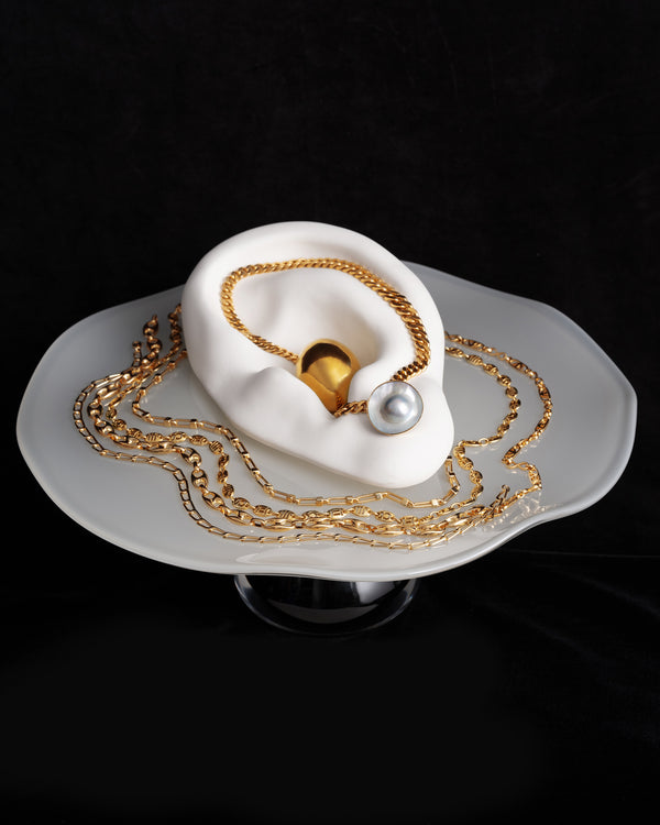 Styled image of various gold chains and pearl earrings on the Bon Bon Cake Stand, on a black background.
