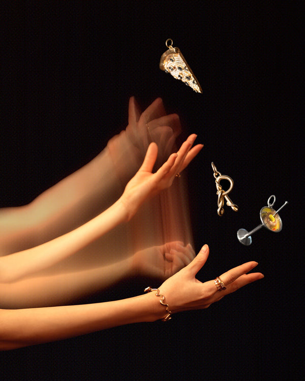 Styled long exposure image featuring the Veggie Slice Pizza Charm, Dirty Martini Charm, Gold Cut-Outs Bracelet and CRZM by Mociun Gold R Letter Charm on dark color background.