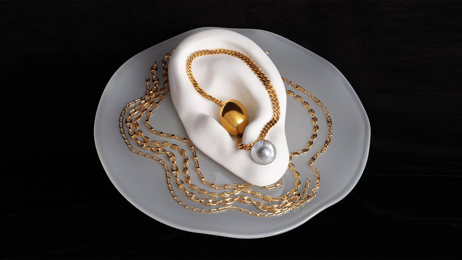 Styled image of various gold chains and pearl earrings on the Bon Bon Cake Stand, on a black background.