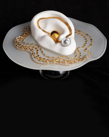 Styled image of various gold chains and pearl earrings on the Bon Bon Cake Stand, on a black background.