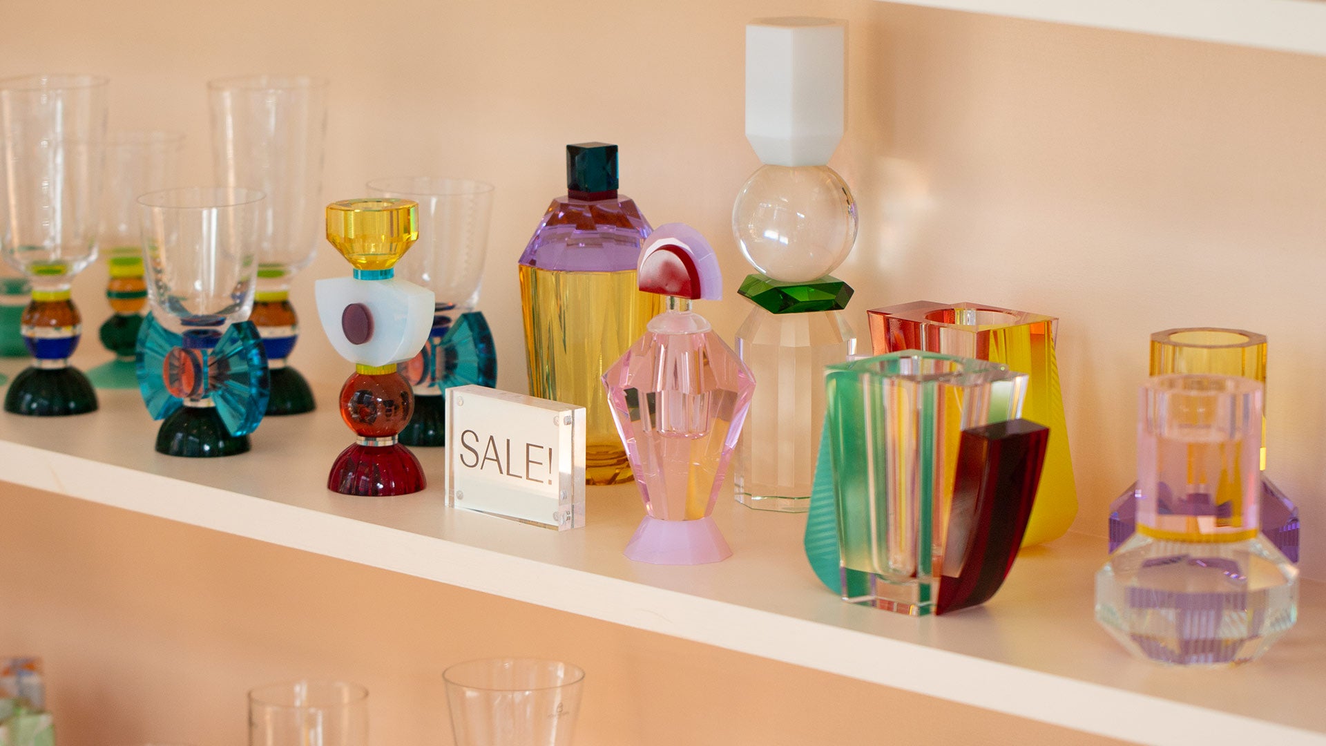 Mociun store interior shelf with "Sale!" sign, showcasing colorful glassware, mixed decor and fine crystal objects.