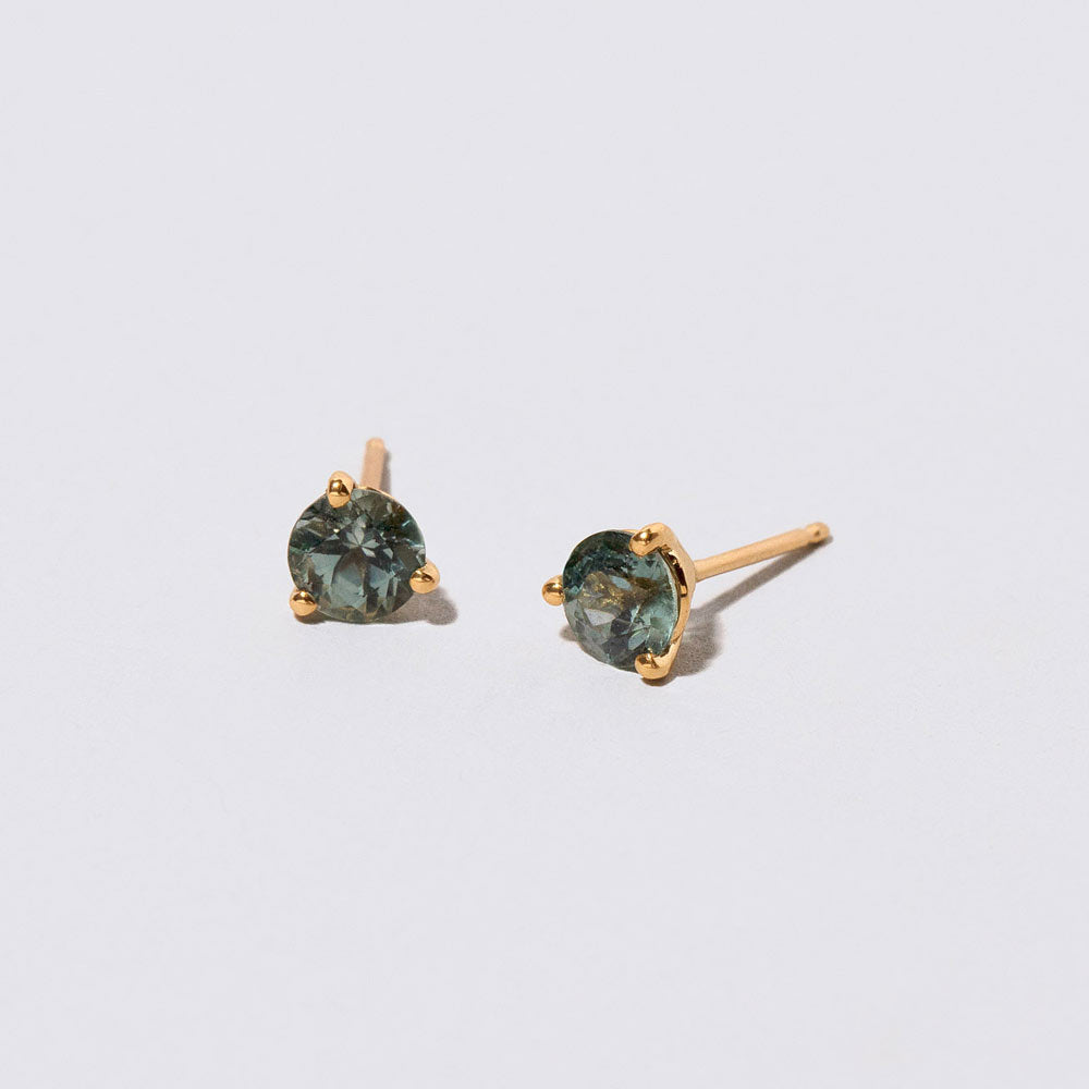 product_details::Closeup details of the Mega Teal Sapphire Martini Stud Earrings on light color background.