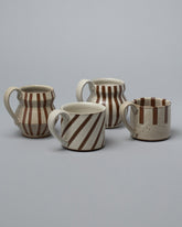 Closeup view of the Jeremy Ayers Mugs Set of Four, including Brown Angle Mug, Brown Diagonal Stripe Mug, Half Stripe Mug, and Brown Bubble Mug on light color background.