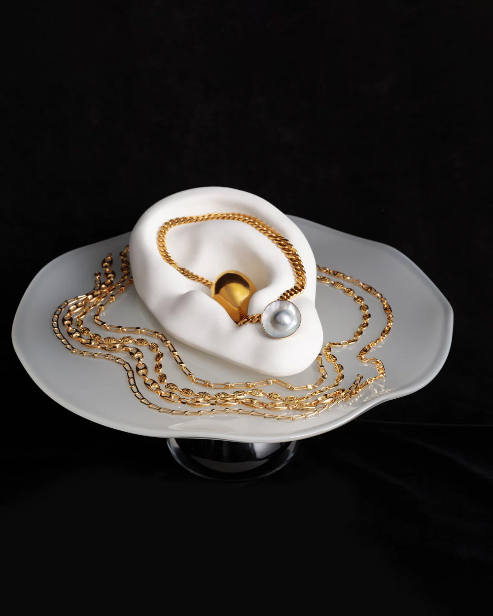 Styled image of various gold chains and pearl earrings on the Bon Bon Cake Stand, on a black background.