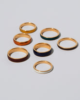 Enamel Bands of various widths and colors on light color background. Photograph represents the 3mm size in Moonbeam (Glow in the Dark) and Earth (Deep brown); the 4mm size in Daffodil (Yellow) and Veridian (Deep Green), the 5mm size in Sunkist (Orange) and Burnt Umber (Deep Red), and in the 6mm size in Cobalt (Navy Blue) and Ink (Black). Edit alt text
