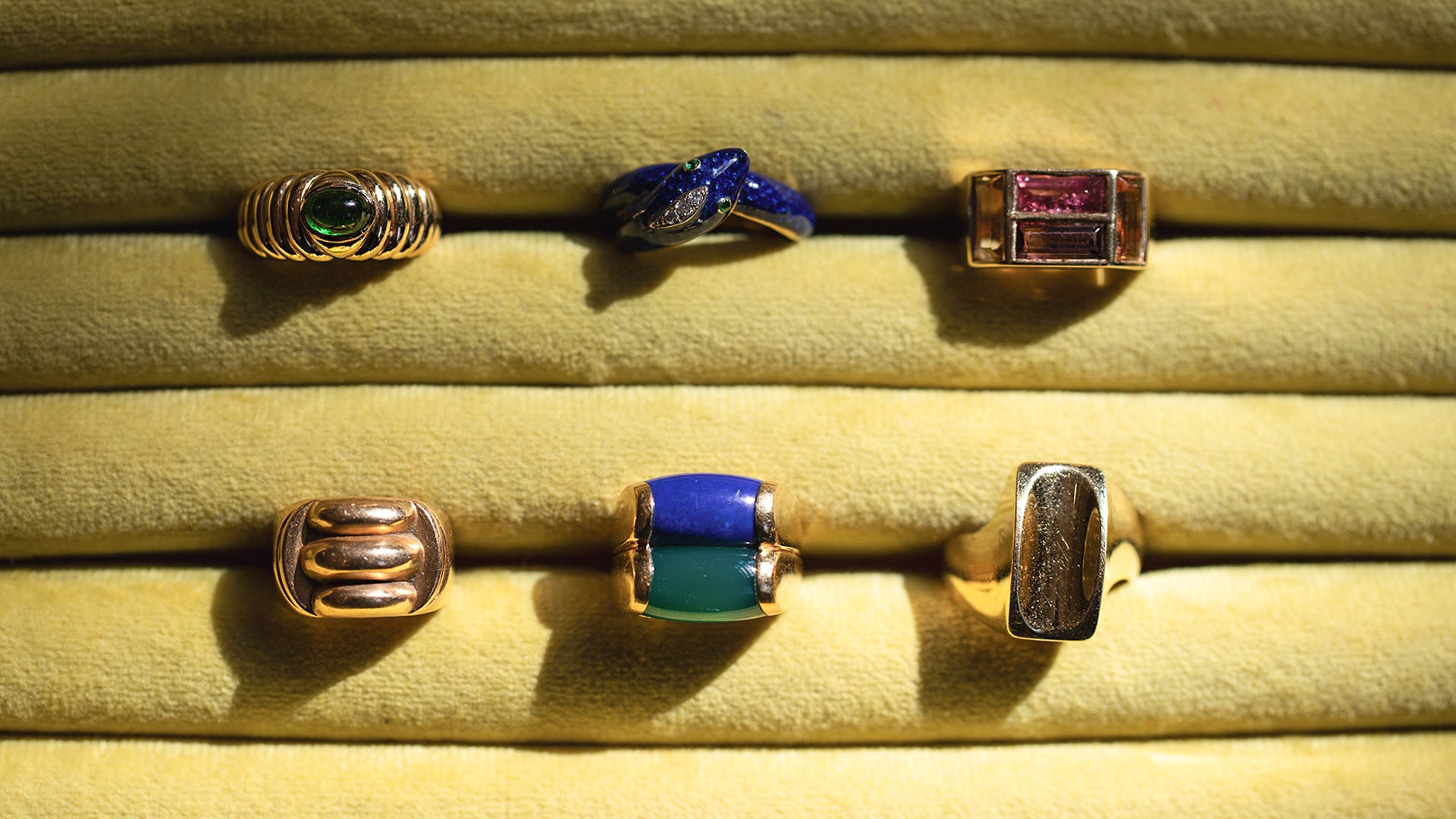 Styled image featuring the Gold Gadroon Ring, Gold Ribbon Ring, Bvlgari Doppio Tronchetto Lapis & Chalcedony Ring, Ribbed Peridot Ring and the Cartier Diamond & Emerald Enamel Snake Ring in green jewelry display.