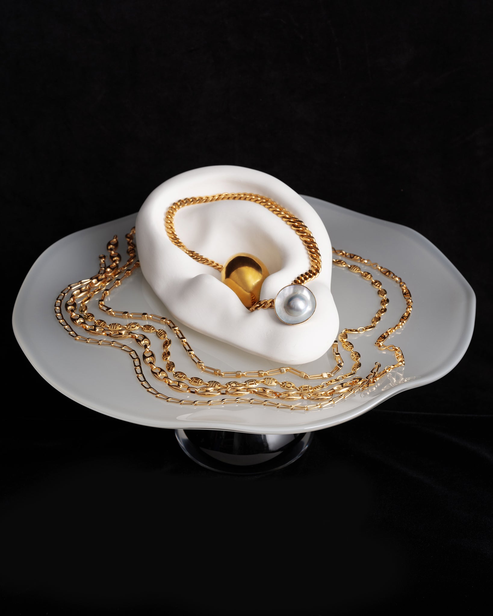 Editorial image of various gold chains and pearl earrings on the Bon Bon Cake Stand, on a black background.