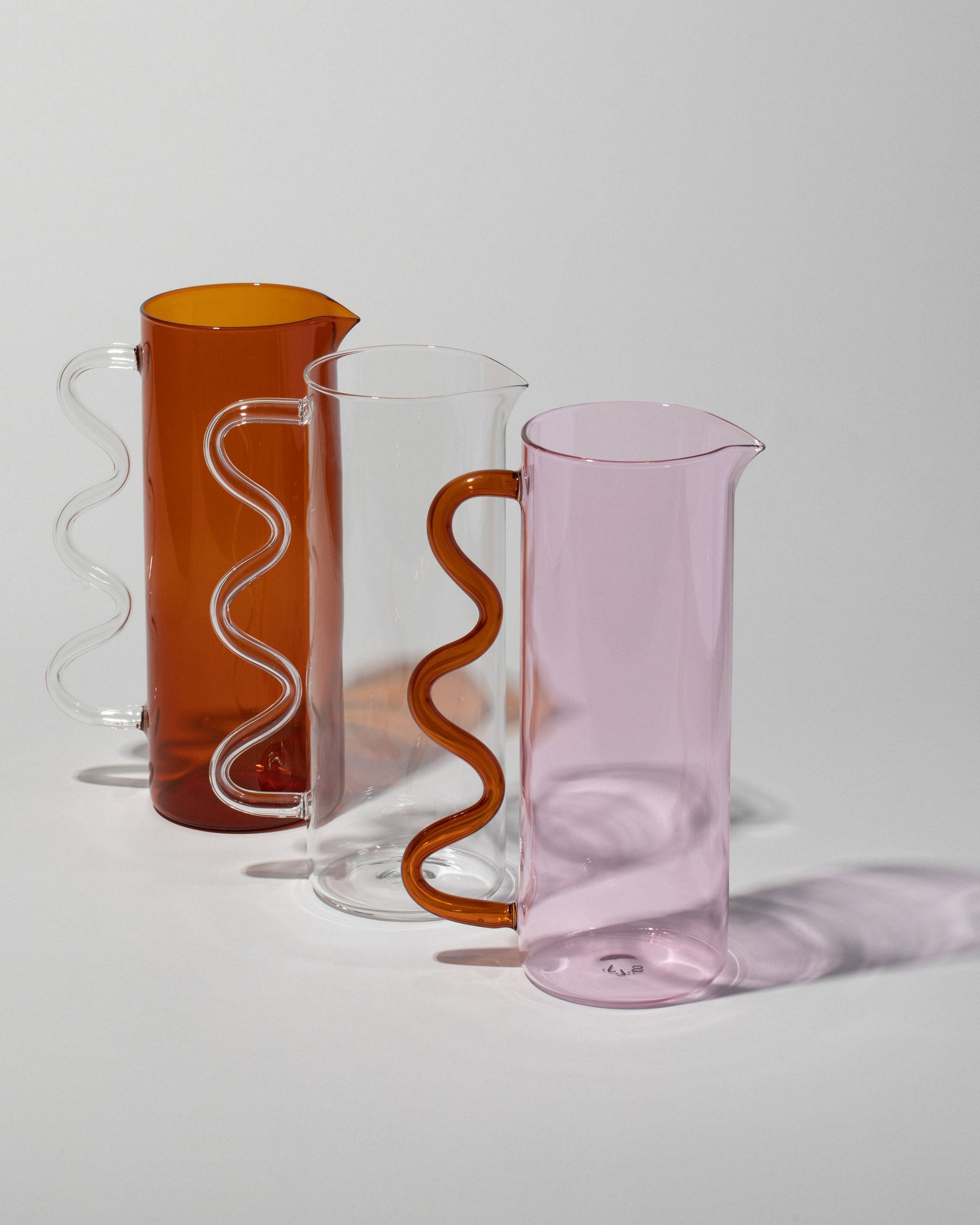 Group of Sophie Lou Jacobsen Clear with Clear Handle, Pink with Amber Handle and Amber with Clear Handle Wave Pitchers on light color background.