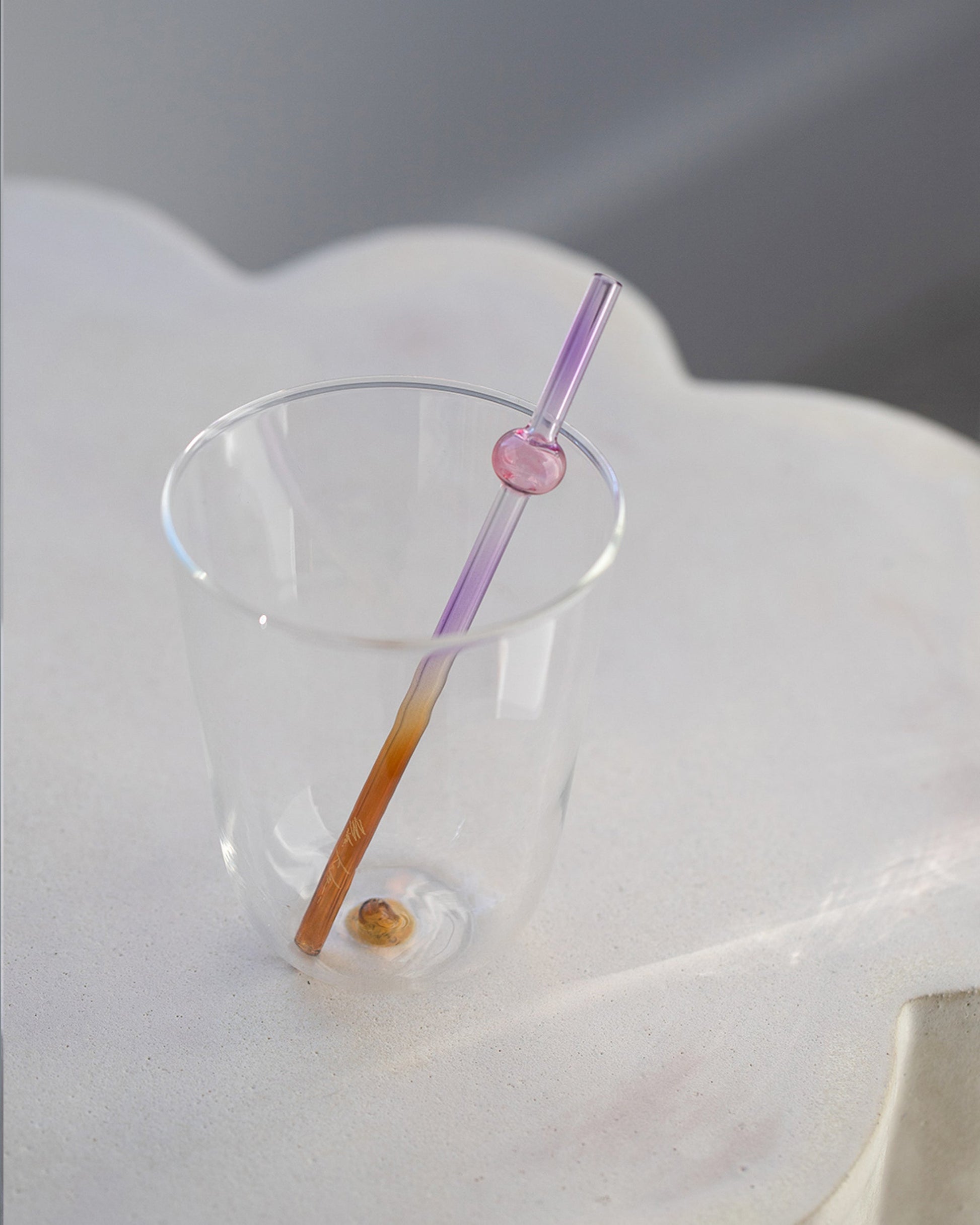 Styled image featuring Misha Kahn Purple and Orange Suck It Up Glass Cocktail Straw and Ichendorf Milano Amber Long Drink Glass on light color background.