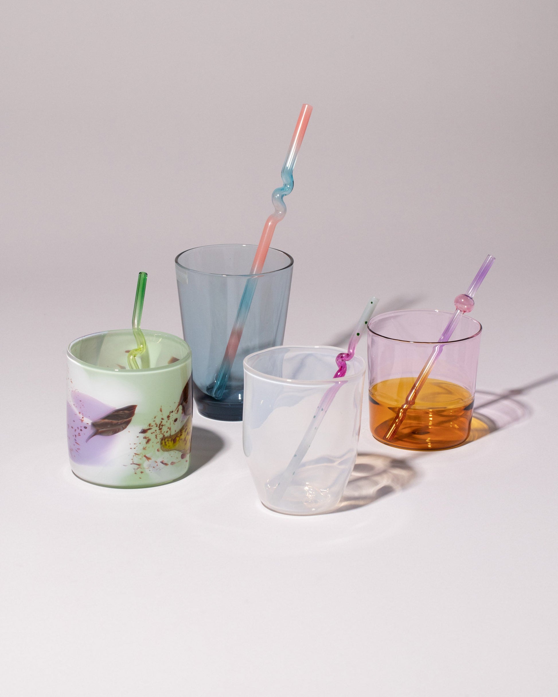 Styled image featuring Misha Kahn Suck It Up Glass Cocktail Straws, Misha Kahn Suck It Up Glass Straws, Balefire Mint Epiphany Cup, Balefire Alabaster Gossamer Cup, Ichendorf Milano Amber/Pink Light Colore Water Glass, and Kinto Hibi Blue Glass on a light colored background.
